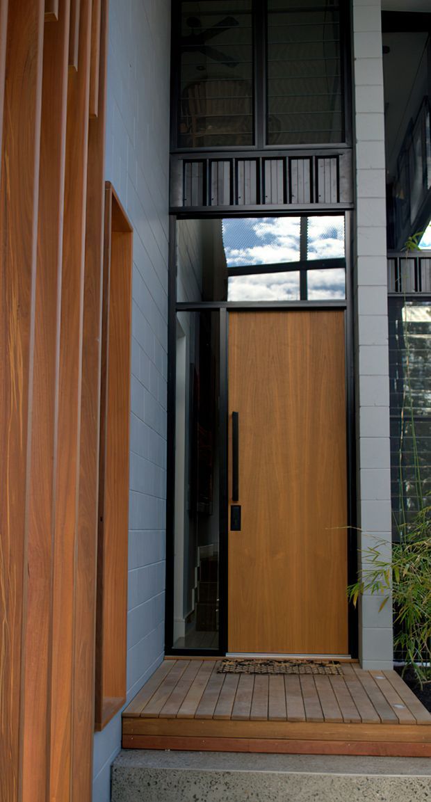 A contemporary residential entryway viewed from outside, featuring a tall timber front door with black hardware, extensive black-framed glazing, a covered double-height entrance, timber wall cladding, pale painted masonry, and a timber deck above a concrete step. Bamboo-like planting is visible at the side.