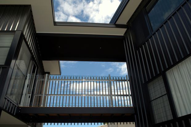 Upward view within a modern multi-level architectural courtyard or atrium, framed by dark metal-clad walls and extensive glazing. A high-level balcony with slim vertical metal balustrades spans the space, while a large overhead skylight reveals blue sky and clouds. The composition emphasises strong geometric lines, structural framing, and contrasting light and shadow.