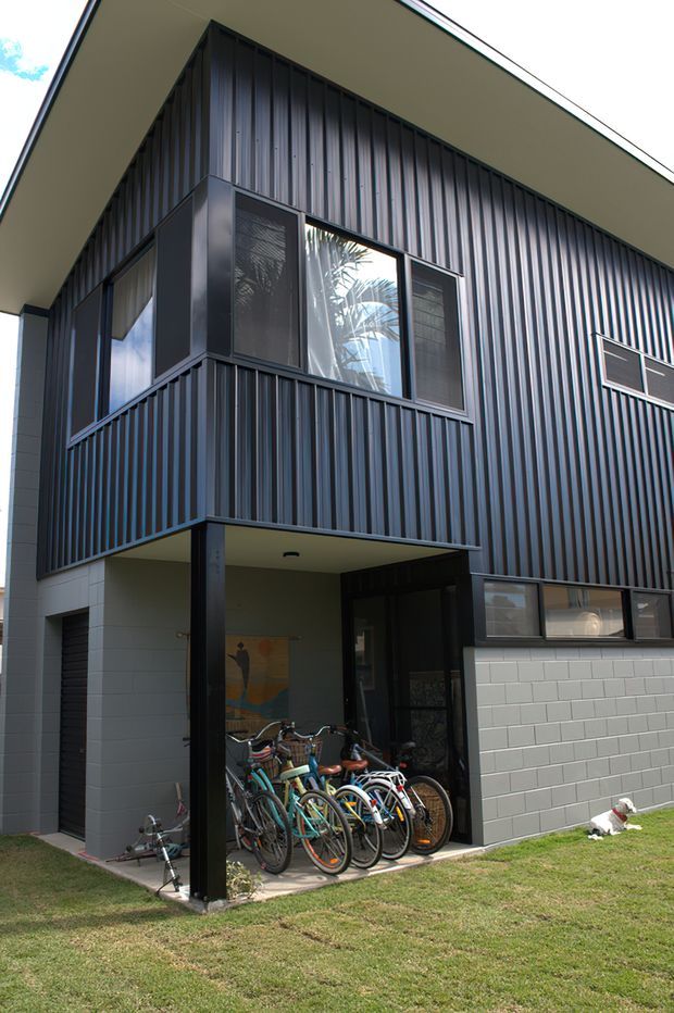 Exterior view of a contemporary multi-storey residential building with dark vertical corrugated metal cladding, projecting upper-level balcony, large windows, and a recessed ground-floor entry. A light masonry wall, garage door, bicycles, and lawn are visible.