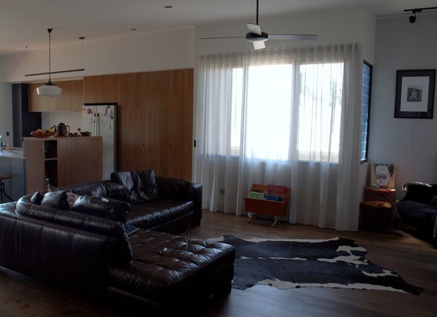 Open-plan living room with a dark leather sectional sofa, tufted ottoman, cowhide rug, timber kitchen cabinetry and matching full-height storage wall. Sheer curtains cover a large window, while ceiling fans and pendant lights provide functional lighting. The space has a warm mid-century-inspired character with a muted, earthy palette.