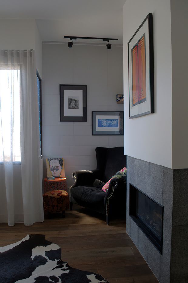 A compact, warmly styled living room corner with a black upholstered armchair, patterned ottoman, framed artwork, sheer curtains, dark timber flooring, and a contemporary built-in fireplace clad in grey stone or tile. Track lighting and layered artwork add a refined, eclectic character.