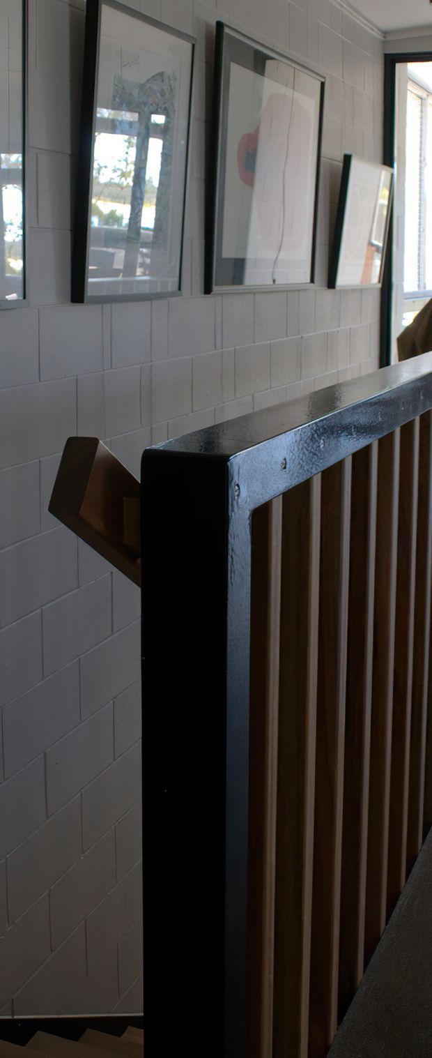 A close-up view of a residential stairway landing with a black-painted railing and vertical timber balusters. The wall is finished in white rectangular masonry or tile blocks and decorated with framed artwork. Natural light enters from a nearby window or glazed opening, creating a contemporary, slightly industrial interior character.