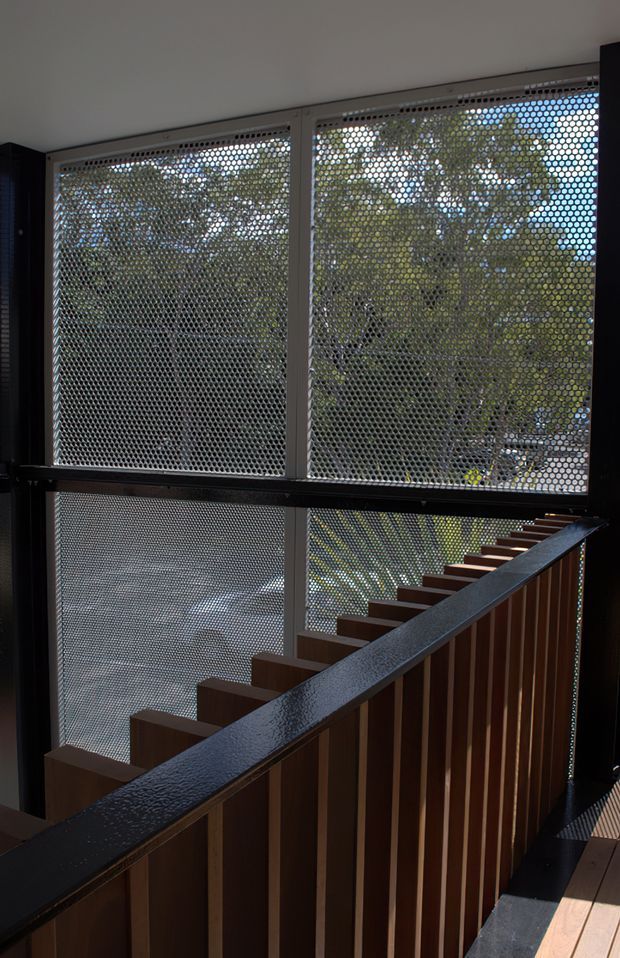 Interior view of a contemporary stairway with dark timber treads and vertical timber balustrade elements, enclosed by large perforated metal screens and framed glazing. Daylight filters through the screens, creating a layered connection to the leafy exterior. The space has a restrained industrial-modern character with strong linear geometry and contrasting dark finishes.