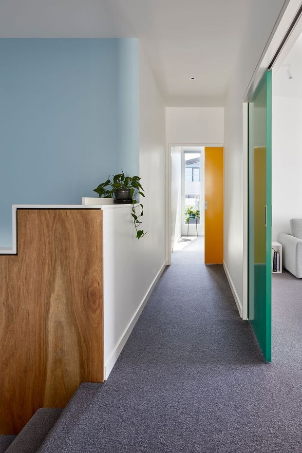 A bright, narrow residential hallway with soft grey carpet, white walls and ceiling, and a pale blue feature wall. A warm timber built-in cabinet with a white top occupies the left foreground, topped by a potted trailing plant. At the far end, a vivid yellow-orange door opens toward a sunlit exterior or balcony. A green-framed glazed or mirrored door on the right reveals part of a light-coloured sofa in an adjoining room. The space has a clean, contemporary and playful colour scheme with strong natural light.