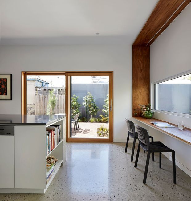 A bright, compact open-plan interior combining a contemporary kitchen with a built-in study nook. White cabinetry and a dark benchtop sit to the left, while a long timber-framed desk with two black chairs runs beneath a horizontal window on the right. Warm timber beams and window frames contrast with white walls and a polished speckled concrete floor. Full-height glazing opens to a small paved courtyard garden.