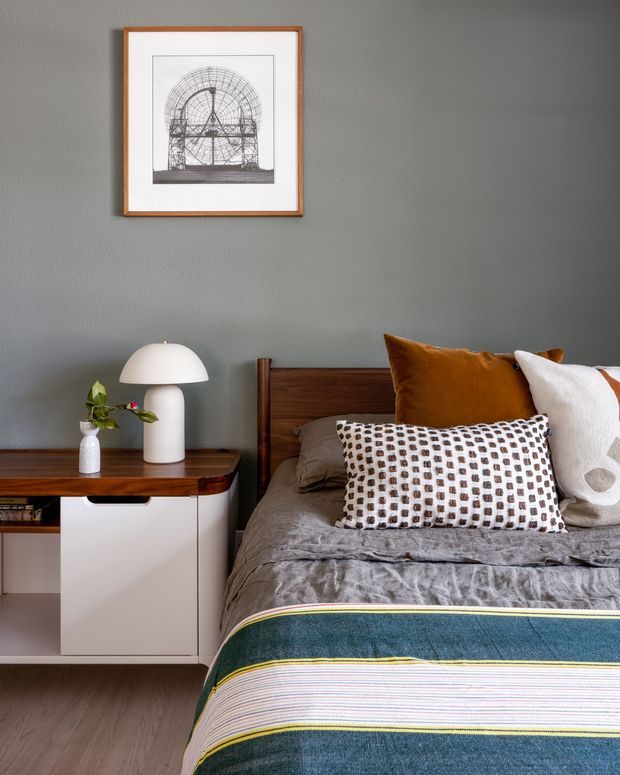 A styled bedroom vignette with a timber bedhead, layered grey bedding, patterned and rust-toned cushions, and a striped blue-green throw. A mid-century-inspired timber and white bedside cabinet holds a white dome table lamp and a small vase. The room has a muted grey painted wall, warm timber flooring, and framed architectural artwork.