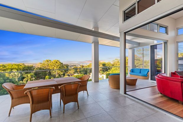 A spacious contemporary outdoor terrace overlooking a leafy suburban landscape. The covered terrace features a tiled floor, glass balustrade, wicker dining furniture, and a large opening connecting to a bright interior living area with vivid blue and red upholstered seating. Floor-to-ceiling glazing, horizontal sun-shading louvers, exposed structural columns, and a high ceiling create an airy indoor-outdoor connection.