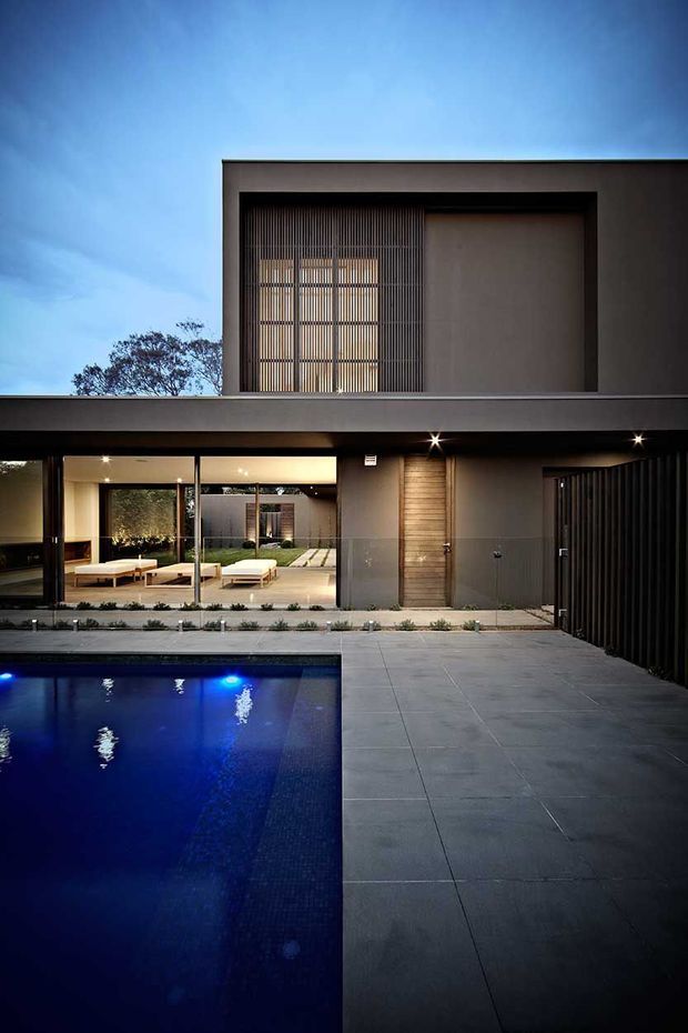 A dusk exterior view of a contemporary two-storey residence organised around a landscaped courtyard and swimming pool. The home has a flat roof, crisp rendered walls, extensive ground-floor glazing, vertical screening, a timber-clad entry door, dark framed openings, and large-format grey paving around the pool. Warm interior and facade lighting contrasts with the blue evening sky and illuminated pool.