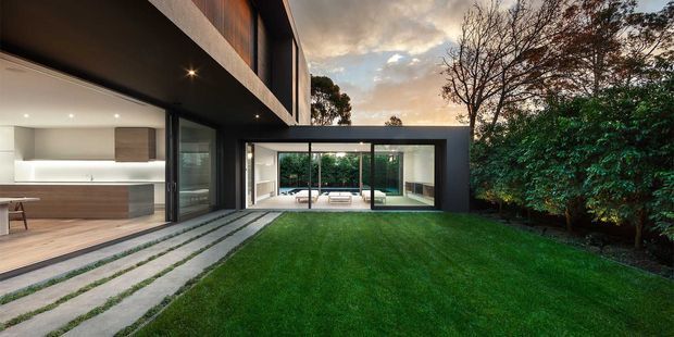 Contemporary two-storey residence viewed from a landscaped lawn and concrete stepping-stone path. The dark, rectilinear exterior frames expansive floor-to-ceiling glazing, with a bright minimalist kitchen visible on the left and a glazed living area opening onto a private courtyard or garden. Strong indoor-outdoor connection, clean geometric forms, and warm timber accents are visible.