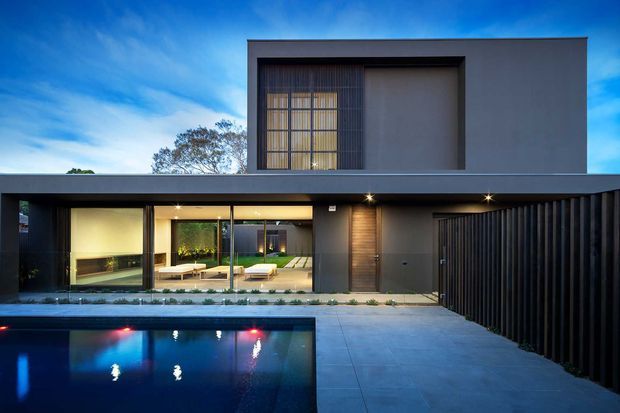 Contemporary two-storey residence viewed from the pool courtyard at dusk. The home has a flat-roofed, rectilinear form with dark charcoal cladding, extensive ground-floor glazing, a tall upper-level window screened by vertical fins, and a broad projecting roof or canopy. A swimming pool, paved forecourt, and dark vertical privacy fence define the exterior setting.