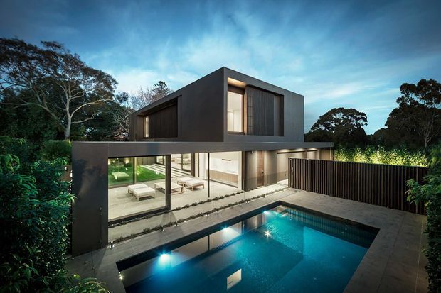 Contemporary two-storey dark-clad residence with large glazed living areas opening to a paved pool terrace. The home features a strong rectilinear form, projecting upper volume, vertical screening, and a rectangular illuminated swimming pool surrounded by established vegetation.