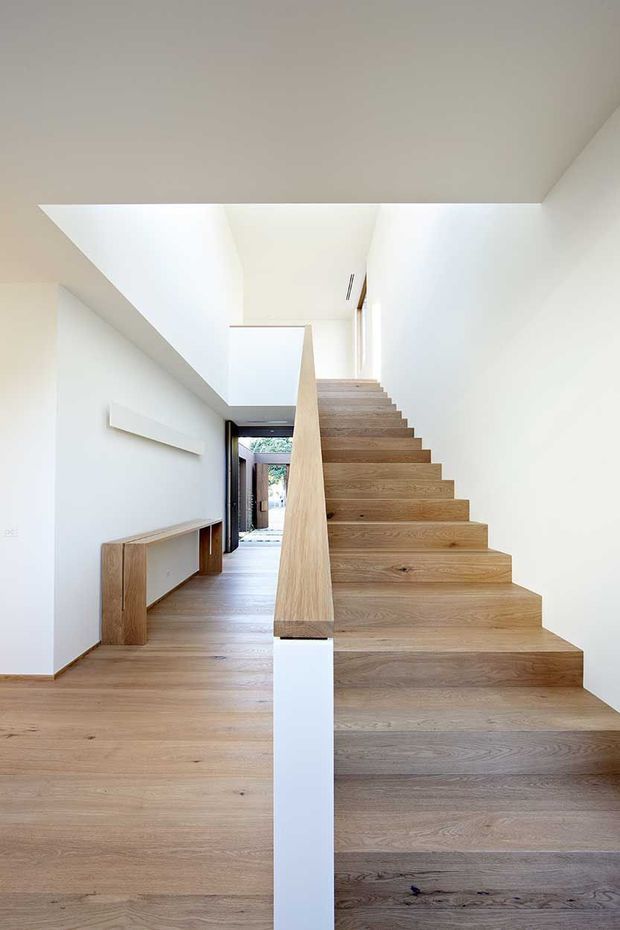 Bright minimalist interior focused on a contemporary timber staircase. Broad oak-toned timber treads continue into the flooring, with a simple white central balustrade and white walls. A small built-in timber console sits along the left wall, while glazing at the rear brings in natural light and provides a view toward an outdoor area.