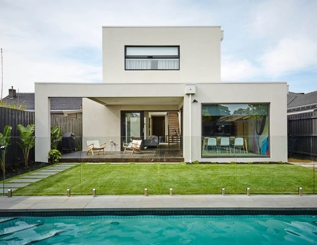 Contemporary two-storey white-rendered home viewed from the backyard, featuring large glazed openings, a covered outdoor entertaining area, timber decking, landscaped lawn, and a swimming pool in the foreground. The rear facade has a simple flat-roofed form with dark-framed windows and sliding doors.