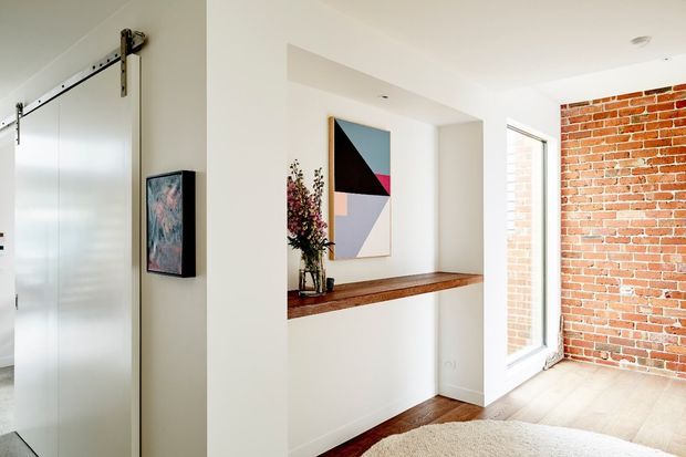 Bright contemporary interior hallway or entry area with a white sliding barn-style door, built-in white wall niche and timber console ledge, abstract artwork, exposed red brick feature wall, and pale timber flooring. Natural light enters through tall glazing, creating a clean, modern space with warm material contrast.
