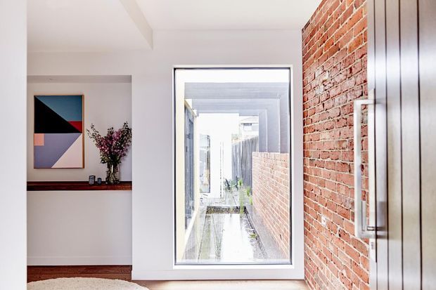 Bright contemporary interior corridor or entry area with an exposed red-brick feature wall, dark vertical timber cabinetry or door panels, white plaster walls and ceiling, and a large black-framed window overlooking a narrow courtyard or passage. A recessed display shelf with abstract artwork and flowers adds a small decorative feature.