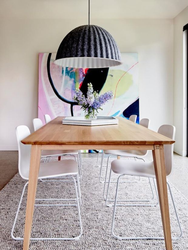 Bright contemporary dining room with a large light timber rectangular dining table, white modern dining chairs, a textured neutral rug, and a prominent black ribbed pendant light. An abstract colourful artwork forms a feature backdrop, with a floral arrangement and stacked books centred on the table.
