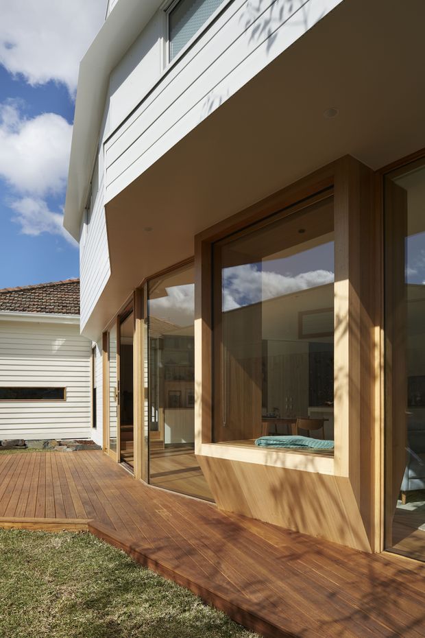 Exterior view of a contemporary two-storey residential extension with white horizontal cladding, broad timber-framed glazing, and a warm timber deck wrapping along the facade. The image also shows an adjoining older single-storey structure with a tiled roof, suggesting a modern addition to an existing home. Strong sunlight creates shadows across the facade and deck.
