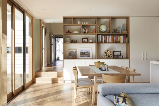 A bright, contemporary open-plan dining and living area with warm timber flooring, full-height timber-framed glazing, and a built-in timber shelving and cabinetry wall displaying books and decorative objects. A timber dining table and chairs sit beside a light upholstered sofa, while a short set of steps leads toward a hallway and the glazed exterior.