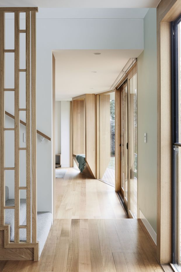 A bright, refined residential hallway or circulation space with pale timber flooring, extensive warm timber wall panelling and joinery, white walls and ceiling, and full-height glazed doors/windows along one side. A timber-framed glazed screen and stair are visible at the left, while a bedroom or living area is glimpsed beyond the corridor. The design emphasizes natural light, clean lines, and integrated timber detailing.