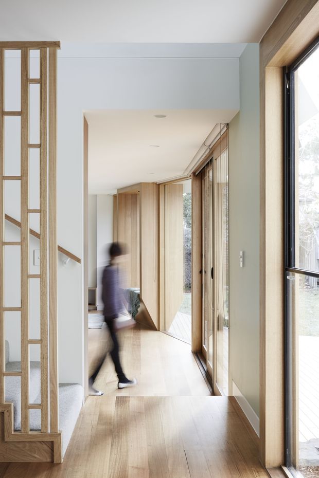 A bright contemporary residential hallway with warm timber flooring, timber-framed glazed doors and windows, and a vertical timber screen partition. The corridor leads toward a living area and an exterior deck or garden visible through extensive glazing. Pale walls, clean lines, and abundant natural light create an airy, calm interior.