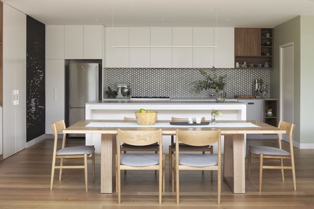 Bright open-plan kitchen and dining area with a long light timber dining table and matching chairs in the foreground. The kitchen features white flat-panel cabinetry, a stainless-steel refrigerator, dark timber shelving and cabinetry, a white island with seating, and a patterned grey-and-white geometric tile splashback. Warm timber flooring and soft neutral wall colours create a clean, contemporary atmosphere.