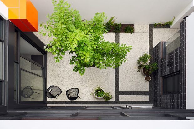Top-down view of a compact contemporary courtyard framed by dark brick and black-framed glazing. A pale terrazzo-like paved surface is arranged in geometric bands around mature trees, raised planters, and potted greenery, with two outdoor chairs and a vivid orange and yellow architectural feature at the upper left.