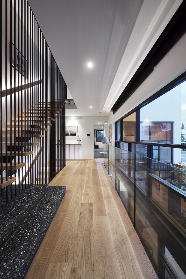 A contemporary upper-level hallway or landing with warm timber flooring, a dramatic open-riser timber staircase enclosed by a black vertical metal balustrade, and extensive dark-framed glazing along one side. The space has white walls and ceiling, recessed lighting, a compact console area, and views into adjacent areas through the glazing.