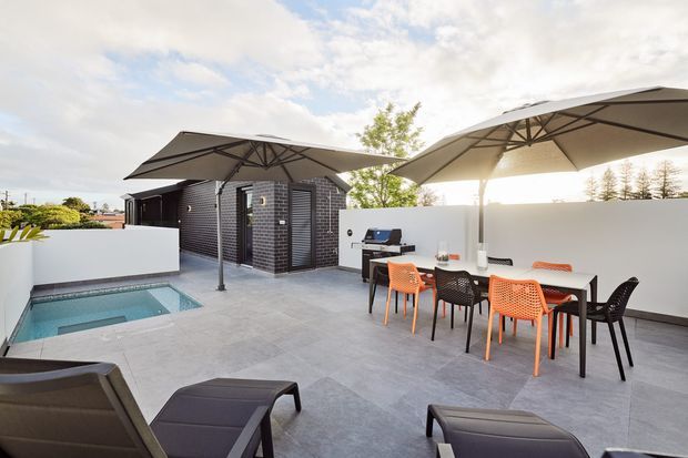 A spacious contemporary rooftop terrace with a small rectangular plunge pool, outdoor dining table and chairs, lounge chairs, large shade umbrellas, and a built-in outdoor barbecue against a dark brick-clad structure. Light grey paving and white rendered walls create a bright, refined outdoor entertaining area.