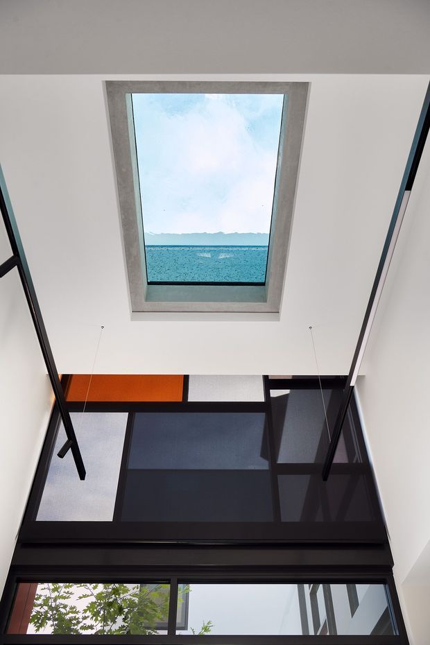 Upward-looking architectural interior view of a contemporary glazed void or atrium. A rectangular skylight with a concrete or cement-look reveal frames the sky and distant water, while large black-framed glazing and dark structural framing create a dramatic double-height space. An orange translucent or coloured panel provides a strong accent near the upper glazing.