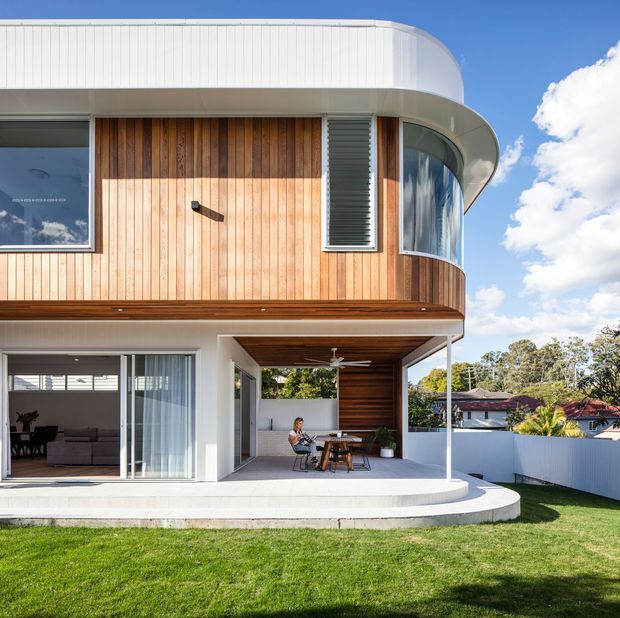 Contemporary two-storey residential home viewed from the rear, featuring curved white upper-level cladding, vertical timber wall cladding, rounded glazing, louvred windows, and a covered outdoor dining terrace with timber-lined ceiling. The house opens onto a lawn and suburban garden.