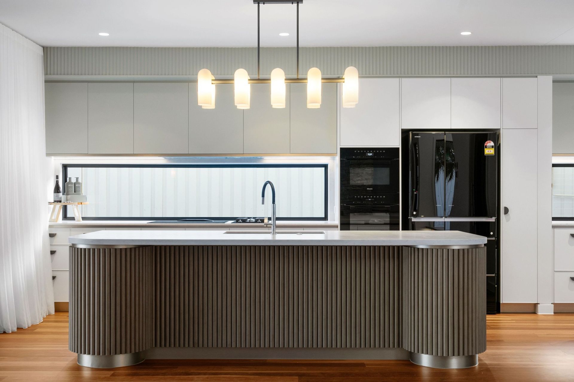 A curved, fluted timber island grounds the kitchen, with a window splashback drawing in garden light.