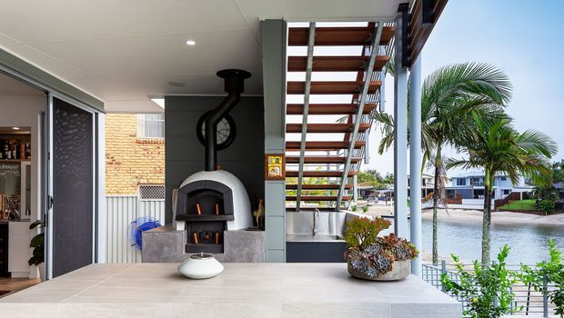 A covered outdoor entertaining area with a tiled counter or island in the foreground, a built-in wood-fired stove, and an open metal staircase leading to an upper level. The space is beside a waterfront or canal, with palm trees and neighbouring buildings visible beyond the glazing. A kitchen or bar area is partially visible to the left.