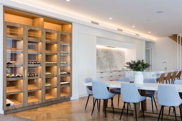 Bright open-plan dining and kitchen interior with a large white dining table, upholstered blue-grey dining chairs, warm timber herringbone flooring, and extensive custom timber-and-glass display cabinetry. A refined white kitchen is visible behind the dining area, including a marble-look feature splashback, island bench, integrated cabinetry, and brass-toned tapware. Recessed ceiling lighting and concealed joinery create a clean, contemporary atmosphere.