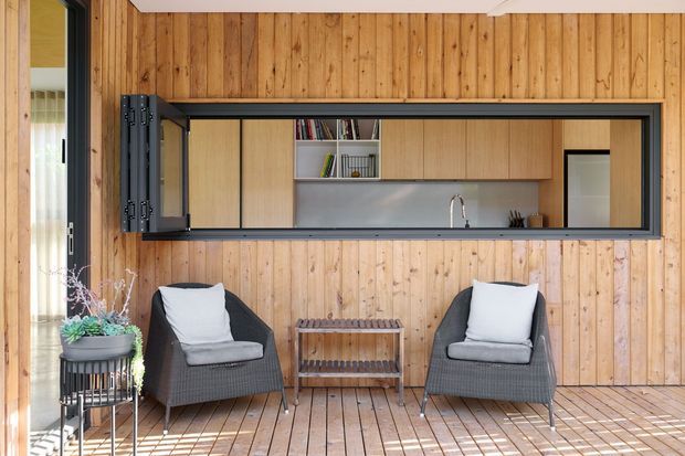 A warm, compact indoor-outdoor living area with vertical timber wall panelling and matching timber decking. Two dark woven lounge chairs with light cushions face a small timber side table. A large black-framed servery-style opening with folding shutters reveals a simple timber kitchen or kitchenette with open shelving, cabinetry, a stainless sink and a pale splashback. Potted plants and strong daylight add a relaxed, natural atmosphere.