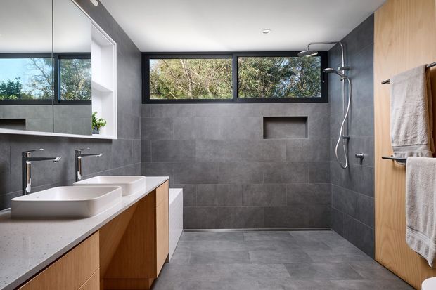 Contemporary bathroom with a walk-in shower, double vessel basins, floating timber vanity cabinetry, large-format grey wall and floor tiles, black-framed clerestory windows, and a timber door with towel rails. The room is bright and clean with views of greenery through the windows.
