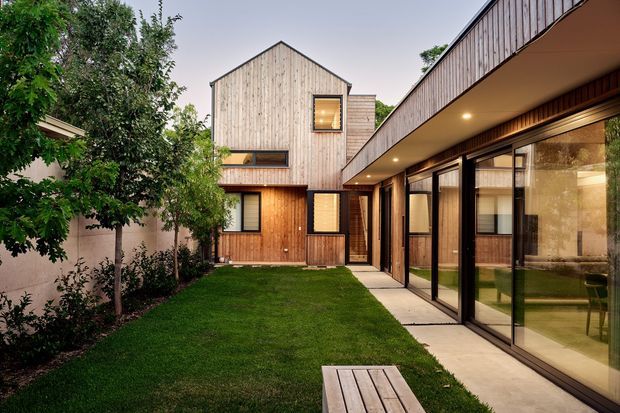 A contemporary courtyard view of a timber-clad residential house. The image shows a compact grass courtyard bordered by a gabled two-storey timber volume and a long glazed single-storey wing with a deep overhanging roof and recessed lighting. Warm interior light, dark-framed glazing, timber decking, and concrete paving create a sheltered indoor-outdoor connection.
