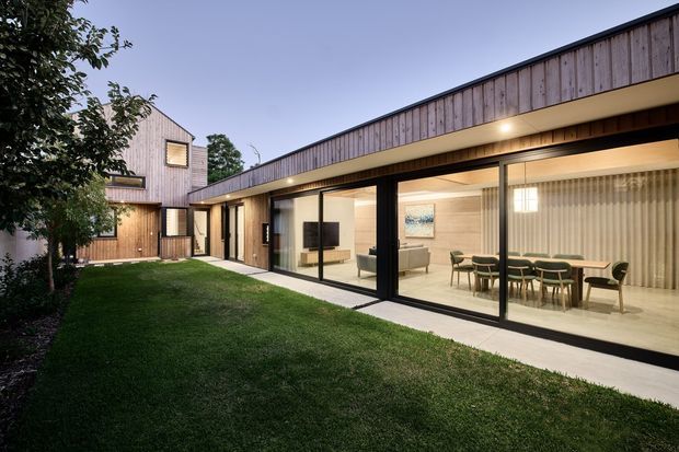A contemporary single-storey residential extension arranged around a landscaped courtyard. The long low pavilion has extensive black-framed floor-to-ceiling glazing, warm timber-clad walls and soffits, and illuminated interior spaces including a living area and dining room. A taller timber-clad volume sits behind the courtyard.