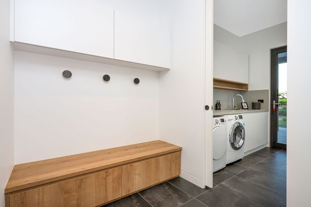 A contemporary laundry and utility area viewed from an adjacent storage or entry zone. The foreground features built-in white overhead cabinetry, a long timber bench with integrated lower cupboards, and wall hooks. Through the opening is a compact laundry with white front-loading appliances, a timber wall shelf, dark tiled flooring, light walls, and a glazed exterior door.