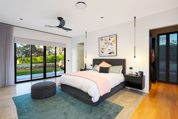 Bright contemporary bedroom with a large upholstered bed, dark bedside tables, pendant lighting, abstract artwork, layered area rugs, and floor-to-ceiling sliding glass doors opening to a landscaped outdoor area. The room combines light neutral walls and bedding with warm timber flooring and blue-green textile accents.