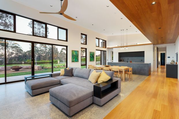 Open-plan contemporary living, dining and kitchen area with expansive black-framed glazing overlooking a landscaped garden. The room features a large grey sectional sofa, timber dining table, dark kitchen cabinetry with a broad island, polished concrete flooring, and a warm timber-lined ceiling and floor zone.