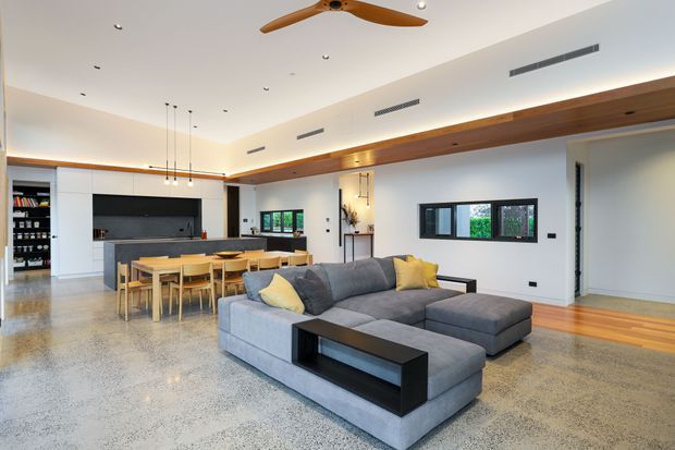 Open-plan contemporary living area with a large grey sectional sofa, black coffee table, timber dining table and chairs, and a streamlined kitchen with island and dark splashback. The room features polished concrete flooring, white walls, extensive timber ceiling beams and a ceiling fan, with windows providing natural light.