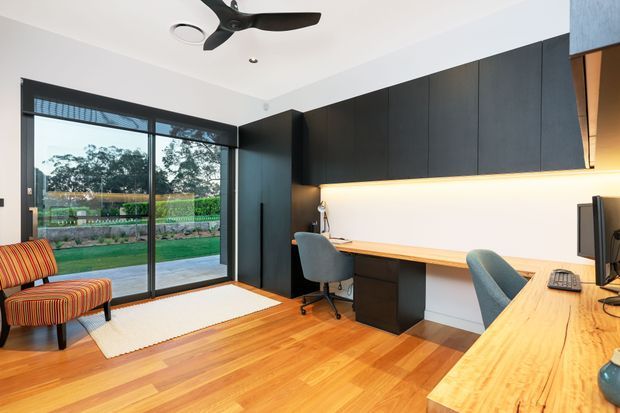 A contemporary home office with extensive built-in dark cabinetry, a long light timber desk and work surface, integrated warm under-cabinet lighting, and two workstations. Floor-to-ceiling black-framed glazing opens views to a garden, while a striped lounge chair, timber flooring, and a black ceiling fan complete the room.