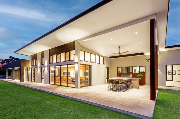 A large contemporary home viewed from the lawn at dusk, featuring broad flat roof overhangs, white rendered walls, dark timber or metal accents, extensive floor-to-ceiling glazing, and a covered outdoor entertaining area. The open-plan space includes a kitchen island with bar seating and a dining area, opening onto a paved terrace and lawn.