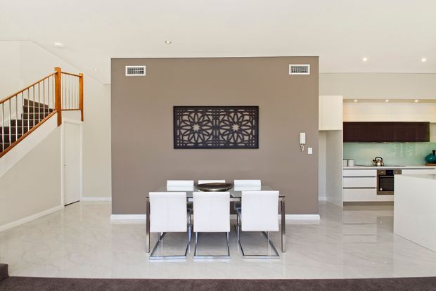 Open-plan interior featuring a contemporary dining area with a rectangular glass-top table and six white upholstered chairs, set against a taupe feature wall with decorative dark artwork. A staircase with timber handrail and metal balustrade is visible to the left, while a minimalist white kitchen is visible in the background. The space has glossy light flooring, white ceilings and cabinetry, and recessed lighting.