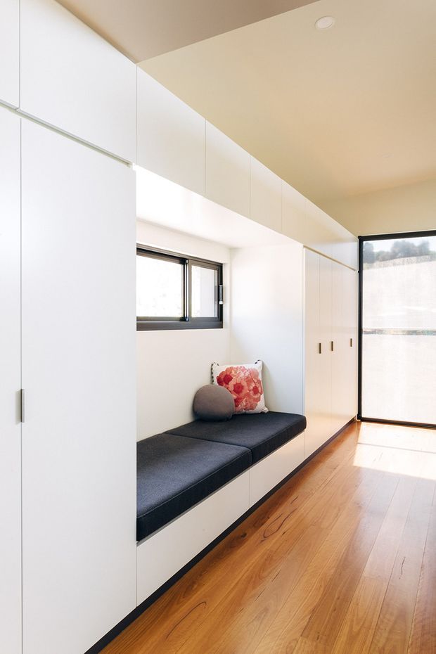 Bright, narrow built-in hallway or entry zone with extensive white floor-to-ceiling cabinetry, a window seat with dark upholstered cushion, timber flooring, and a large glazed door or window at the far end. The space has clean modern joinery, warm beige ceiling tones, and strong natural light.
