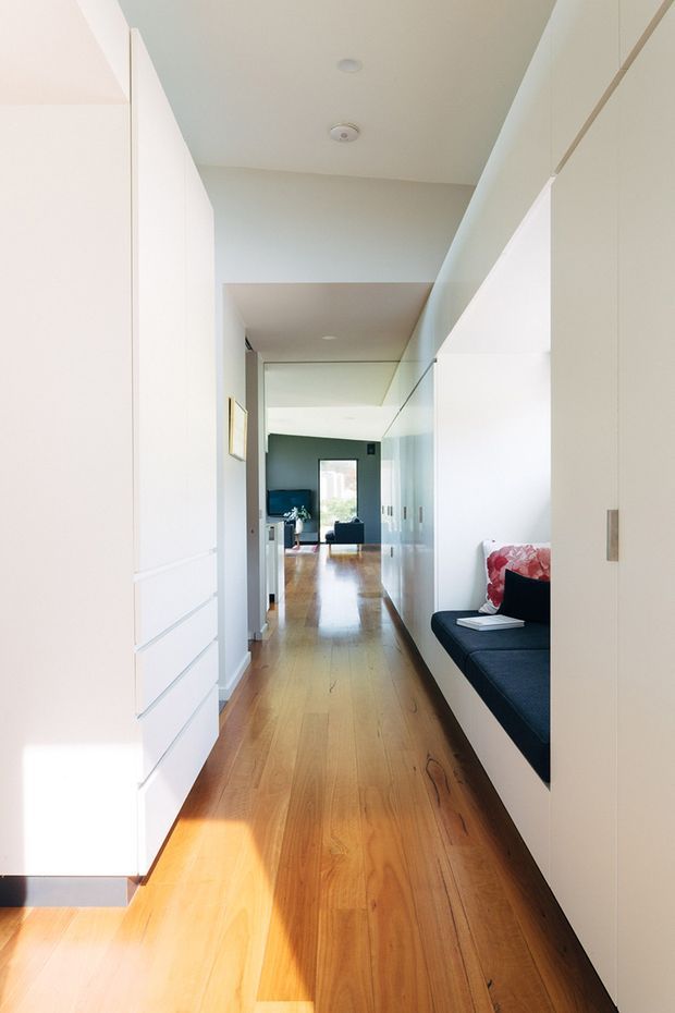 A bright, narrow residential hallway with full-height white built-in cabinetry on both sides, a glossy timber floor, and a partially enclosed built-in seating nook visible on the right. The corridor leads toward a living area with a dark accent wall, lounge chair, and large window. The overall design is clean and highly minimalist, with strong natural light and concealed storage.