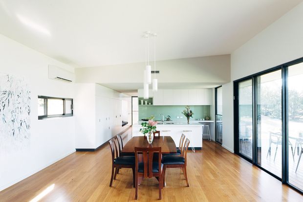 Bright open-plan dining and kitchen interior with warm timber flooring, a large dark wood dining table and chairs, white cabinetry, a pale green tiled splashback, and extensive black-framed glazing opening toward an outdoor area. Minimal pendant lights and clean-lined finishes create an airy contemporary feel.