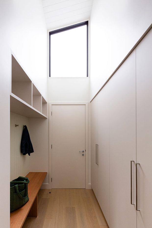 A narrow, bright built-in storage corridor or entry area with full-height white cabinetry on the right, open shelving and a timber bench on the left, and a simple white door at the end. Warm timber flooring and bench seating contrast with the white walls and joinery. A tall upper window and white slatted ceiling bring in abundant natural light, while dark metal window framing and cabinet handles provide subtle contrast.
