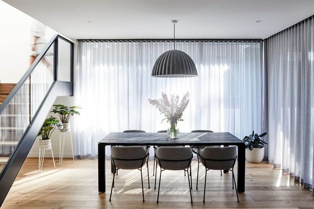 Bright contemporary dining area with a dark rectangular dining table and six upholstered chairs, positioned in front of full-height sheer curtains. A large ribbed dome pendant hangs above the table, while a glazed stair enclosure and indoor plants add architectural and natural elements. Light timber flooring and abundant filtered daylight create an airy, refined atmosphere.