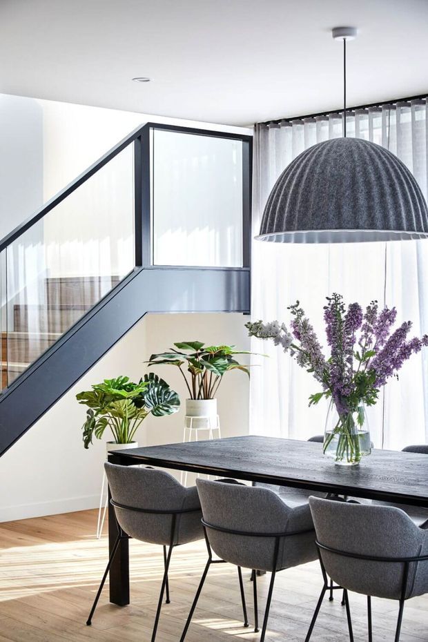 Bright contemporary dining area with a long dark dining table and upholstered grey chairs, positioned beside a glazed staircase with dark framing. Sheer curtains, indoor plants, a floral arrangement and a large ribbed pendant light add texture and softness. Light timber flooring and white walls create an airy, refined interior.